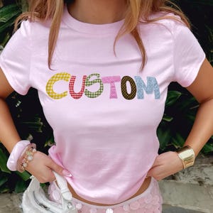 Op de afbeelding: Een lichtroze t-shirt met het woord "CUSTOM" in kleurrijke patchwork letters. Het model draagt een bijpassende roze pailletten minirok, een witte handtas en gouden sieraden.