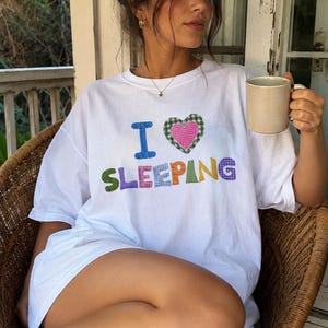 May include: A white short-sleeved t-shirt featuring the text I LOVE SLEEPING in colorful, patterned fabric letters. The letter O is replaced by a heart shape with a pink center and a green checkered border. The letters feature various patterns.