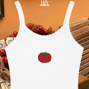 Pu&ograve; includere: Canotta bianca con spalline sottili e grafica di pomodoro rosso. La canotta ha uno scollo tondo e una silhouette aderente. La grafica del pomodoro &egrave; ricamata con foglie verdi. La parola "LAZY" &egrave; stampata in alto.