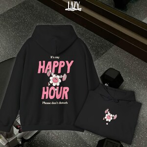 May include: Black hoodie with the text "It's my HAPPY HOUR" in pink, featuring a flower lifting weights. The phrase "Please don't disturb" is below. A second hoodie with the same design is also visible.