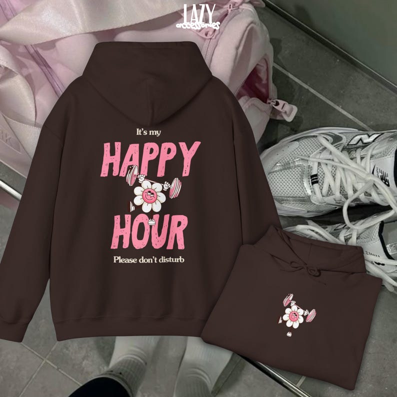 May include: A brown hoodie with the text "It's my HAPPY HOUR Please don't disturb" in pink. The design includes a white flower with weights. A second folded hoodie with the same design is also visible.
