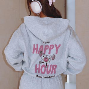 May include: A light gray hoodie with the text "It's my HAPPY HOUR" in pink, featuring a cartoon flower lifting weights. The phrase "Please don't disturb" is printed below. The hoodie is part of a matching set.