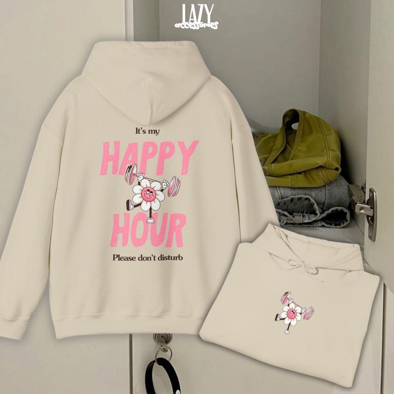 May include: Cream-colored hoodie with a pink and white graphic of a flower lifting weights, and the text "It's my HAPPY HOUR. Please don't disturb." A second hoodie is folded below. The word "LAZY accessories" is at the top.
