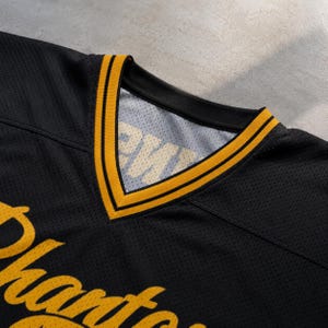 May include: A black mesh jersey featuring a V-neck collar with yellow and black striped trim. The front of the jersey displays yellow cursive lettering, and the interior neck label shows partial text reading PHANTOMS in a bold font.