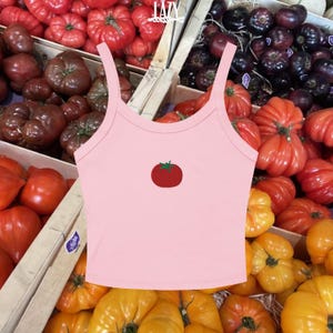 Pu&ograve; includere: Top corto rosa chiaro con grafica di pomodoro rosso. Il top ha spalline sottili e scollatura rotonda. Lo sfondo presenta vari tipi di pomodori in casse di legno.