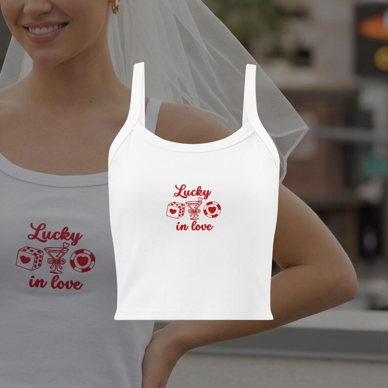 May include: White tank top with the phrase "Lucky in love" in red script. The design includes a die, a martini glass, and a casino chip. The top has a scoop neck and spaghetti straps.