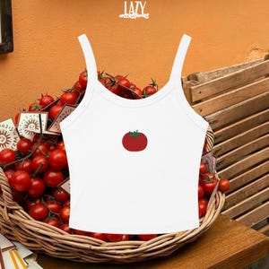 Pu&ograve; includere: Canotta bianca con spalline sottili con grafica di pomodoro rosso. La canotta &egrave; esposta davanti a un cesto di pomodori rossi. La parola "LAZY" &egrave; stampata nella parte superiore dell'immagine.