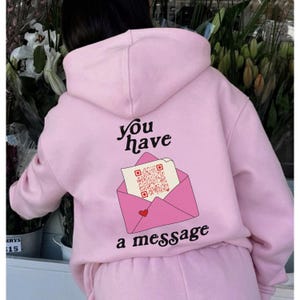 Könnte beinhalten: Rosa Hoodie mit einem Aufdruck eines rosa Umschlags mit einem Herz und einem QR-Code. Der Text "you have a message" ist auf dem Hoodie aufgedruckt.