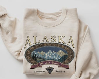 May include: A cream-coloured sweatshirt with the word "ALASKA" in tan lettering. The design features an oval illustration of mountains, trees, and wildlife, with the text "The Last Frontier" and "American Tradition".