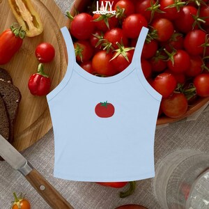 Pu&ograve; includere: Un top corto azzurro con grafica di pomodoro. Il top ha spalline sottili e scollatura rotonda. L'immagine mostra anche un tagliere in legno con pomodori e un coltello.