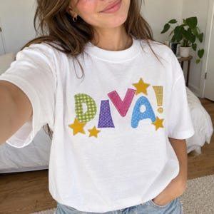 Może przedstawiać: Biały t-shirt z krótkim rękawem z napisem DIVA! wykonanym z kolorowych, wzorzystych liter z tkaniny. Litera D jest w zieloną kratkę, I w fioletowe kropki, V jest różowa, a A w niebieskie kropki. Całość zdobią żółte gwiazdki i wykrzyknik.