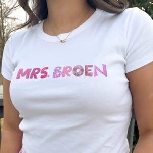 Puede incluir: Camiseta blanca con el texto "MRS. BROEN" en letras rosas de patchwork. La camiseta tiene cuello redondo y mangas cortas. Se ve un collar dorado con pequeños dijes.