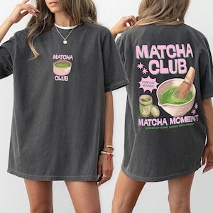 Puede incluir: Camiseta gris oscuro con temática de matcha. La parte delantera presenta un gráfico de tazón de matcha con el texto "Matcha Club". La parte trasera muestra un diseño más grande con un tazón de matcha, un batidor y las palabras "Matcha Club" y "Matcha Moment".