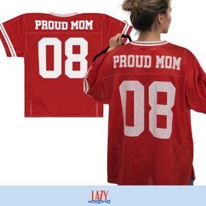May include: A red mesh jersey featuring white text that reads PROUD MOM and the number 08 in a bold athletic font. The garment has white striped detailing on the sleeves and collar, presented in both a flat view and a worn view.