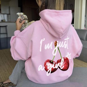 Może przedstawiać: Różowa bluza z kapturem z białym nadrukiem dwóch wiśni i tekstem "I'm just a girl".