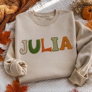 Op de afbeelding: Een beige sweater met ronde hals en de naam JULIA in grote, kleurrijke letters van bedrukte stof. Elke letter heeft een eigen ontwerp, waaronder groene ruitjes, oranje stippen en strepen, allemaal voorzien van zichtbare stiksels.