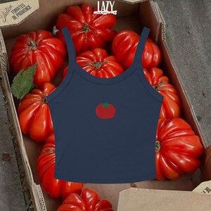 Pu&ograve; includere: Un top corto blu navy con una grafica di pomodoro rosso. Il top ha spalline sottili e uno scollo tondo. Lo sfondo presenta una scatola di cartone piena di grandi pomodori rossi.