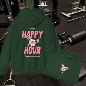 May include: Dark green hoodie with the text "It's my HAPPY HOUR" in pink, featuring a flower graphic lifting weights. The phrase "Please don't disturb" is below. A second hoodie is folded below the main one.