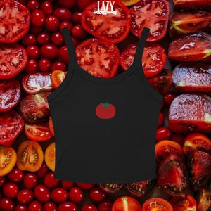 Pu&ograve; includere: Canotta nera con grafica di pomodoro rosso. La canotta ha scollo tondo e spalline sottili. Lo sfondo &egrave; una collezione di pomodori rossi, alcuni tagliati.