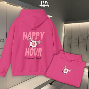 May include: Bright pink hoodie with the text "It's my HAPPY HOUR" and a cartoon flower lifting weights. The phrase "Please don't disturb" is below the text. A second folded hoodie is in the lower right corner.