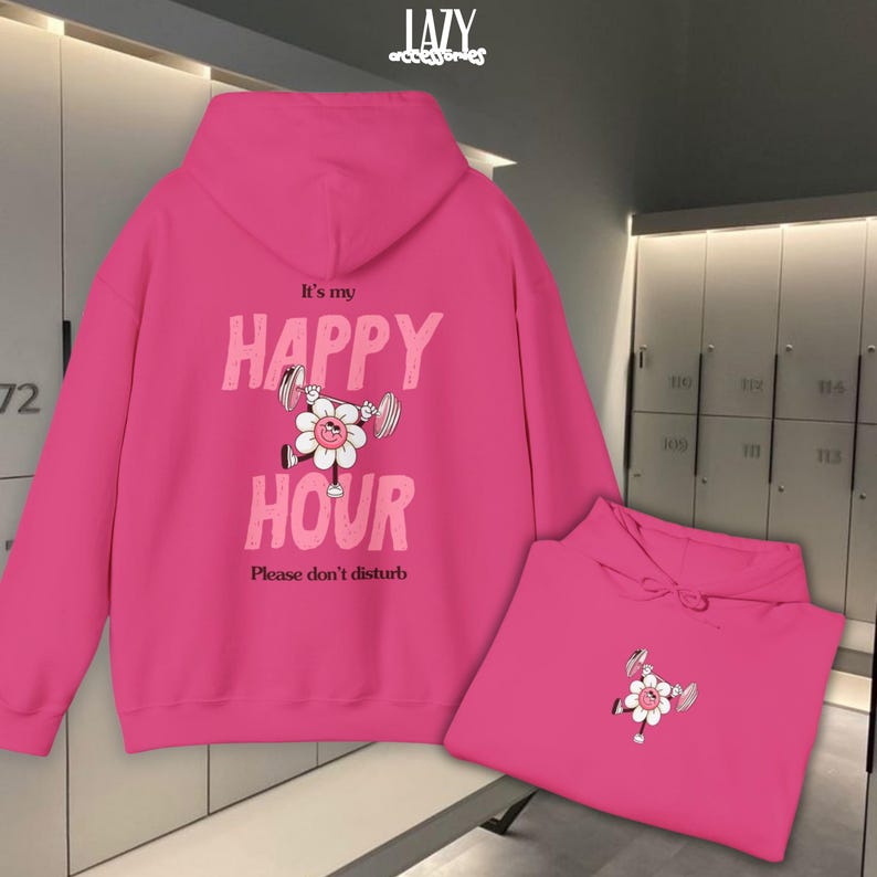 May include: Bright pink hoodie with the text "It's my HAPPY HOUR" and a cartoon flower lifting weights. The phrase "Please don't disturb" is below the text. A second folded hoodie is in the lower right corner.
