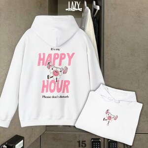 May include: White hoodie with the text "It's my HAPPY HOUR" in pink, featuring a flower lifting weights. The phrase "Please don't disturb" is below. A second folded hoodie with the same design is also visible.