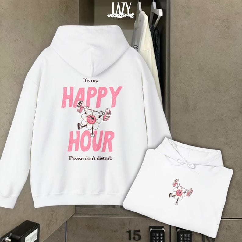May include: White hoodie with the text "It's my HAPPY HOUR" in pink, featuring a flower lifting weights. The phrase "Please don't disturb" is below. A second folded hoodie with the same design is also visible.