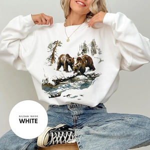 May include: White sweatshirt with a graphic of two brown bears in a snowy scene. The top has a crew neck and long sleeves. The text "GILDAN 18000 WHITE" is printed on a white circle.