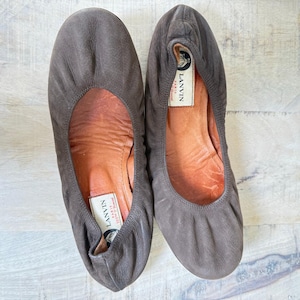 Puede incluir: Un par de bailarinas de cuero marrón. Los zapatos tienen una punta redondeada y un diseño fruncido. El interior del zapato es de color marrón claro. El nombre de la marca "LANVIN PARIS" es visible en la plantilla.