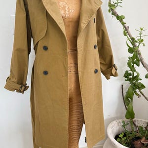 May include: A tan trench coat with a double-breasted design, featuring large lapels, a belt, and button details. The coat is displayed on a wooden dress form. The word "WOLF" is visible on the dress form.