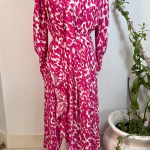 May include: A long, flowing dress in a vibrant pink and white spotted pattern. The dress features long sleeves, a high neckline with a bow, and a ruffled detail on the front. The dress is displayed on a vintage dress form.