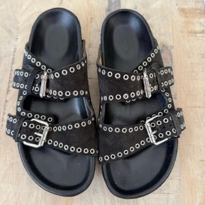 May include: A pair of black suede sandals with silver-tone metal buckles and eyelet details. The sandals have two adjustable straps and a contoured footbed. The soles are black.