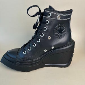 May include: Black leather high-top sneaker with a wedge heel. The shoe features a lace-up design with silver eyelets, a rubber toe cap, and the Converse All Star logo on the side. The heel is approximately 4 inches.