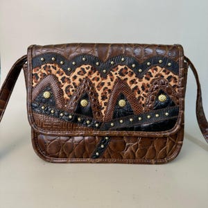 May include: A brown leather shoulder bag with a patchwork design featuring leopard print, crocodile print, and black leather. The bag has gold-toned studs and accents.