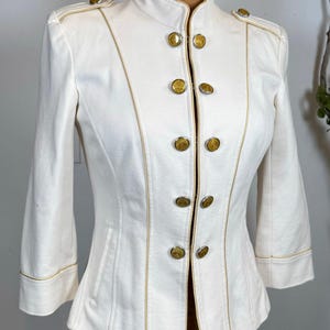 Puede incluir: Chaqueta color crema con ribetes y botones dorados. La chaqueta presenta un cuello alto y mangas tres cuartos. Los botones son dorados y están alineados en la parte delantera de la chaqueta.