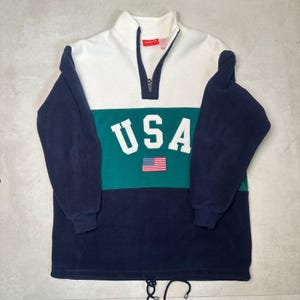 May include: A white, green, and navy blue fleece pullover with a half-zip closure. The pullover has the word "USA" in white letters on the front with a small American flag.