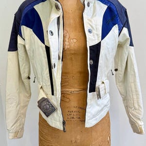 May include: A vintage ski jacket in cream, navy blue, and black. The jacket features a high collar, a zip-up front, and a decorative silver buckle. The jacket is displayed on a vintage dress form. The jacket has a fitted waist and a sporty design.
