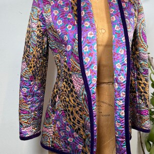 May include: A quilted jacket with a vibrant paisley and floral pattern in shades of purple, teal, and gold. The jacket has a dark purple velvet trim and is displayed on a vintage dress form.