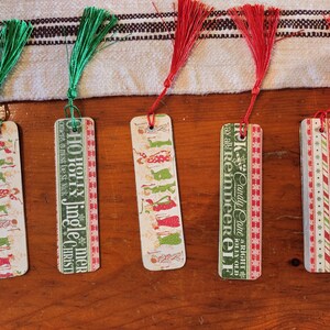 Op de afbeelding: Vijf kerstthema-bladwijzers met rode en groene kwastjes. De bladwijzers bevatten illustraties van de kerstman, rendieren en snoepstokken. De tekst op de bladwijzers luidt "HO HO HOLLY Jingle CHIRST", "Candy Cane a RIGHT JOLLY OLD Vay all! ReinDEER ELF", en "DYS #TOYS #TOYS TO".