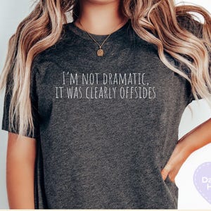 May include: Dark gray heather t-shirt with the text "I'M NOT DRAMATIC, IT WAS CLEARLY OFFSIDES" in white. The shirt is a casual style, suitable for everyday wear.