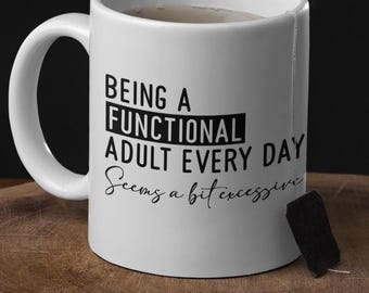 Könnte beinhalten: Weiße Keramik-Tasse mit dem Text "Being a Functional Adult Every Day Seems a bit excessive" in Schwarz. Die Tasse steht auf einem Holzschneidebrett mit einem Holzlöffel.