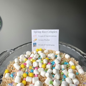 Puede incluir: Un cuenco de vidrio lleno de Spring Rice Crispies. El postre incluye malvaviscos, Rice Krispies y chispas de colores. Una tarjeta de receta enumera los ingredientes: 5 tazas de malvaviscos, ¼ taza de mantequilla y 6 tazas de Rice Krispies.