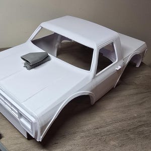 May include: White unpainted plastic model car body for a truck. The body has a scoop on the hood and a rear window.