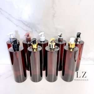 Könnte beinhalten: Acht bernsteinfarbene Glasflaschen mit goldenen und silbernen Pumpenaufsätzen. Die Flaschen sind in einer Reihe auf einer weißen Marmorfläche angeordnet. Der Text "LZ WHOLESALE" ist in der rechten unteren Ecke des Bildes sichtbar.