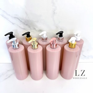 Könnte beinhalten: Acht rosa zylindrische Flaschen mit verschiedenen farbigen Pumpenaufsätzen. Die Aufsätze sind schwarz, gold, weiß, silber, roségold und schwarz. Die Flaschen sind in einer Reihe auf einer weißen Marmorfläche angeordnet. Der Text "LZ WHOLESALE" ist in der rechten unteren Ecke des Bildes sichtbar.