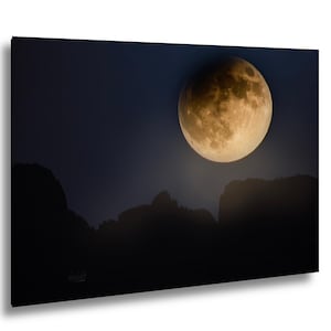 May include: A rectangular wall art print featuring a large, golden-hued moon partially illuminated against a dark blue night sky. Below the moon, the dark, jagged silhouettes of mountain peaks are visible against the horizon in the lower section.