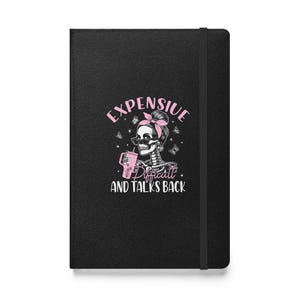 May include: Black notebook featuring a pink and white graphic of a skeleton with sunglasses and a pink bow. The text reads "Expensive and Talks Back". The notebook has a black elastic band.