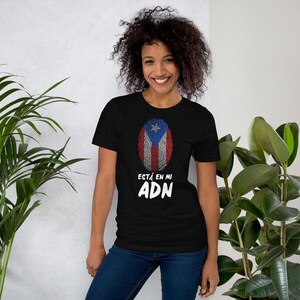 Puede incluir: Una camiseta negra con una impresión blanca de una huella dactilar con la bandera puertorriqueña en su interior. El texto "ESTÁ EN MI ADN" está impreso debajo de la huella dactilar.