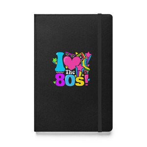 May include: Black notebook featuring the phrase "I love the 80s!" in vibrant colours. The design incorporates a pink heart, stars, and retro graphics. The notebook includes a black elastic band closure.
