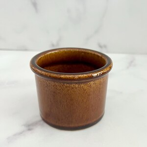 Pode incluir: Um pequeno recipiente cilíndrico de cerâmica castanha com uma borda ligeiramente alargada. O exterior tem um aspeto texturizado e salpicado, e o interior é de um tom castanho mais escuro. O recipiente é provavelmente uma pequena tigela ou prato.